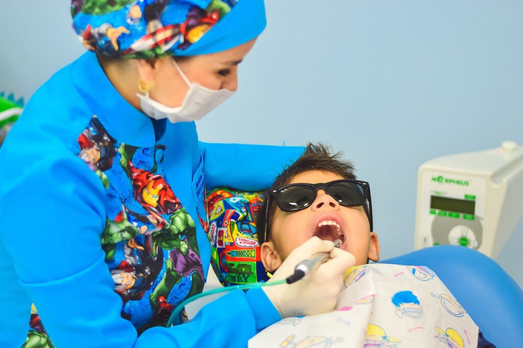 A woman in a blue shirt and hat assists a child seated in a dentist chair during a dental cleaning appointment.