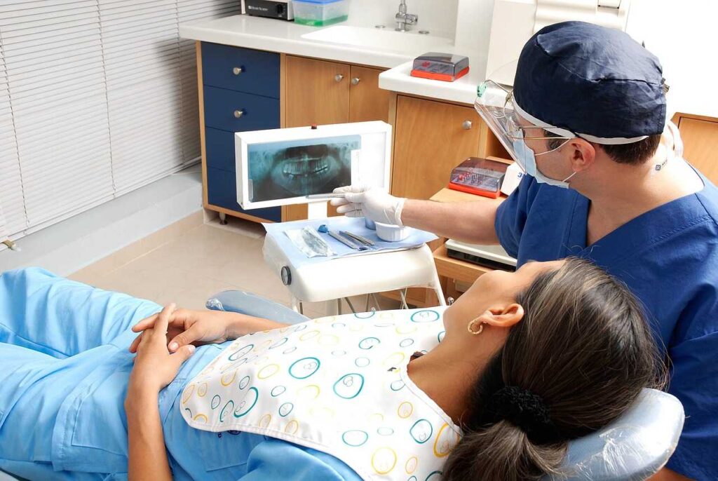 In an emergency dentistry setting, a woman is in a dental chair with a man visible in the background.